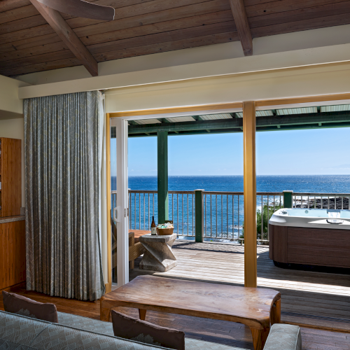 A cozy indoor living space opens to a scenic ocean view with a hot tub on the deck outside, blending comfort with natural beauty.