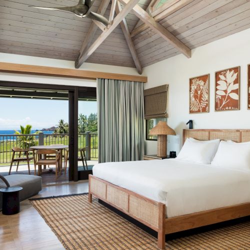 A stylish hotel room featuring a large bed, wooden decor, and a seating area with an ocean view through large windows and a balcony door.