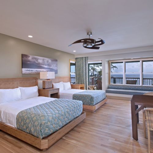 A spacious hotel room with two double beds, a seating area, a desk, and a large window offering an ocean view. Modern decor with wooden accents.