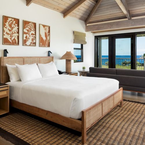 A modern bedroom features a white bed, wooden furnishings, woven lamps, wall art, a grey sofa, and large windows open to a view of the ocean.