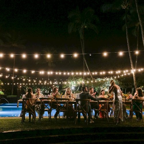 Outdoor evening gathering by a pool with string lights overhead; guests seated at tables eating and socializing.