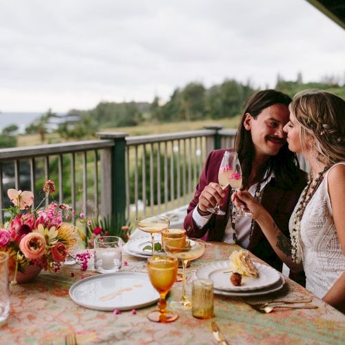 A couple is enjoying a romantic dinner on a balcony with a scenic view, sharing a toast with glasses of wine, and smiling at each other.