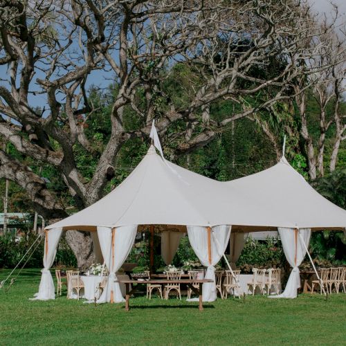 A white tent set up on a grassy area with wooden tables and chairs underneath, surrounded by lush greenery and trees on a cloudy day.