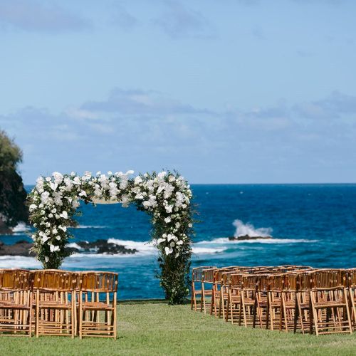 An outdoor wedding setup with rows of wooden chairs and a floral arch sits on a grassy area by the ocean with a scenic rocky coastline in the background.