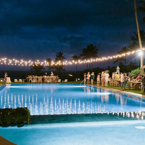 A nighttime outdoor gathering by a pool with string lights overhead, palm trees in the background, and people socializing.