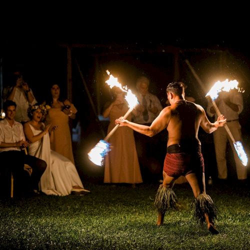 A fire performer entertains an audience during an outdoor event at night, with people seated and standing while watching the act with lit torches.