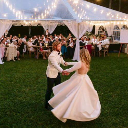 A couple dances under a white tent decorated with string lights, surrounded by guests seated at tables, during an outdoor evening event.