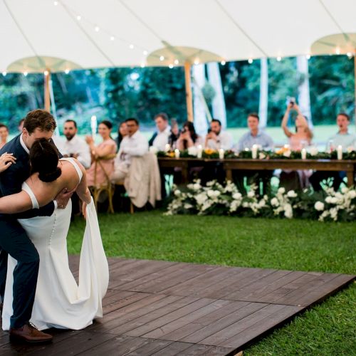 A couple dances under a white tent with guests seated and watching them. There is greenery and floral decorations around the area, and string lights above.