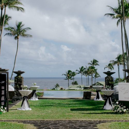 A beautiful outdoor event setup with tables, decorations, and a pathway leading towards the ocean, surrounded by tall palm trees under a cloudy sky.