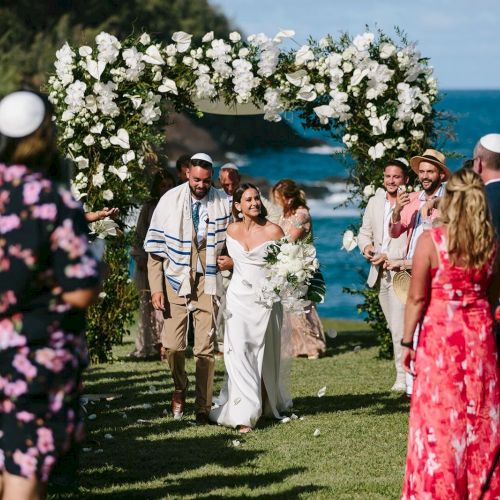 A wedding ceremony by the ocean, with a couple walking down the aisle under a floral arch, surrounded by guests in festive attire.