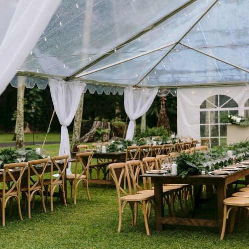 A wedding or event setup with wooden tables and chairs under a transparent tent, adorned with greenery and white drapes, on a grassy lawn.