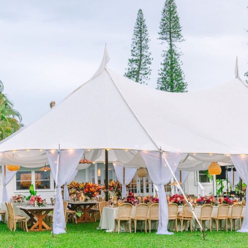 An elegant outdoor event setup under a large white tent with draped curtains, tables and chairs arranged, and lush greenery in the background ending the sentence.