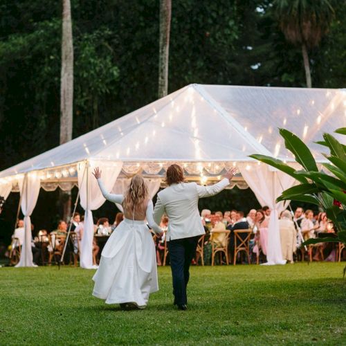 A couple is walking towards a beautifully lit tent, possibly at an outdoor event or wedding, with guests seated inside the tent.