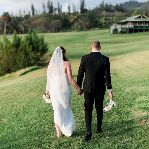 A bride and groom walk hand in hand across a grassy field, with the bride holding a bouquet and the groom carrying his shoes.