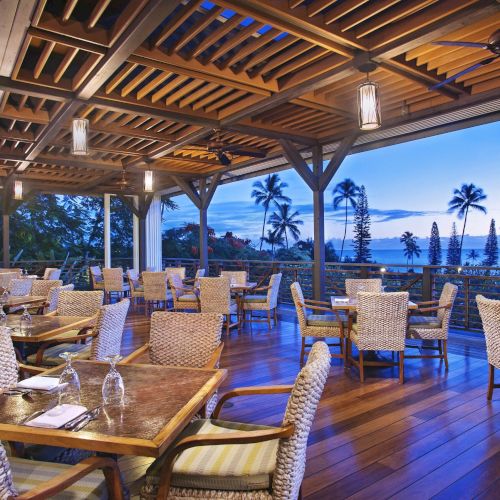 Outdoor restaurant with woven chairs and wooden tables, overlooking palm trees and the ocean at sunset. The area features a wooden pergola ceiling.