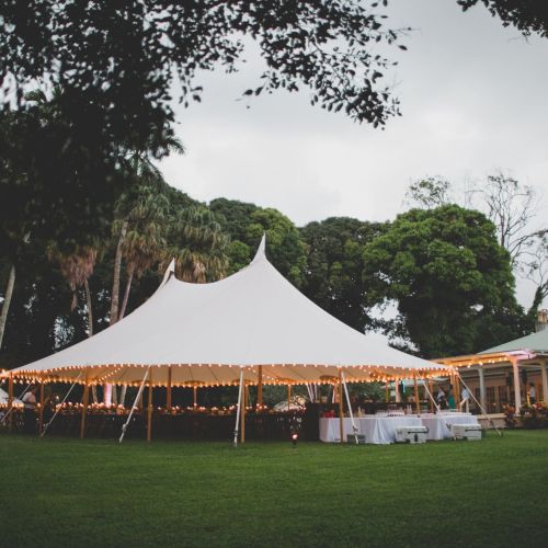 A picturesque outdoor event with a large white tent, string lights, and surrounded by trees and greenery, creating a charming atmosphere.