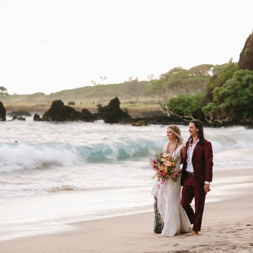 A couple in wedding attire walks hand-in-hand along a scenic beach with waves crashing and rocky cliffs in the background.