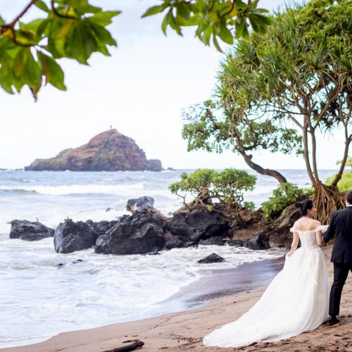 A bride and groom walk on a scenic beach with rocky outcrops and greenery, gazing toward the ocean and a distant small island.