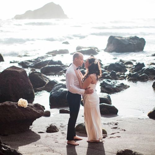 A couple is standing close together on a rocky beach by the ocean, with a bouquet placed on a rock nearby.