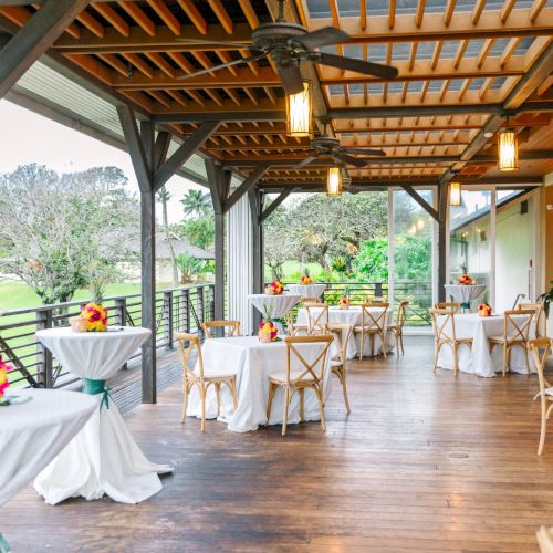 An outdoor dining area with wooden furniture, tables set with colorful floral arrangements, and warm lighting from ceiling fans and lanterns.