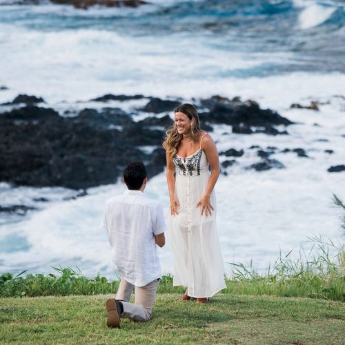 A person is kneeling, proposing to another beside an ocean with waves crashing on rocks in the background, both are smiling and happy.