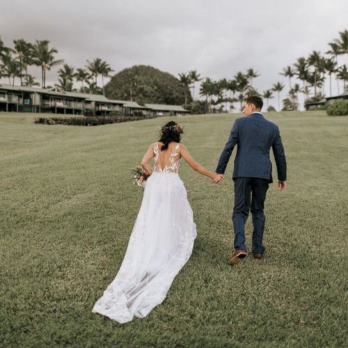 A couple in wedding attire, holding hands on a grassy field with palm trees and buildings in the background, walking away from the camera.