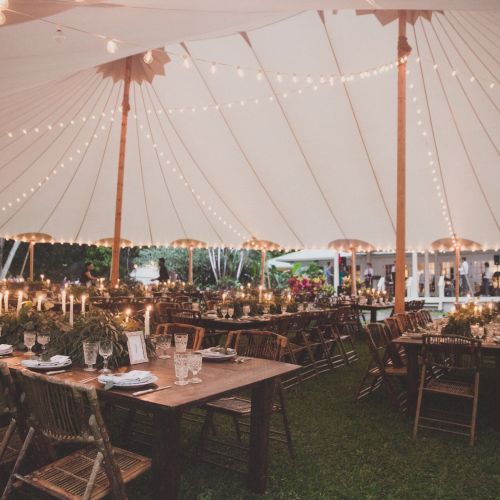 The image shows an outdoor event under a large white tent with string lights, featuring decorated tables and wooden chairs, likely for a celebration.