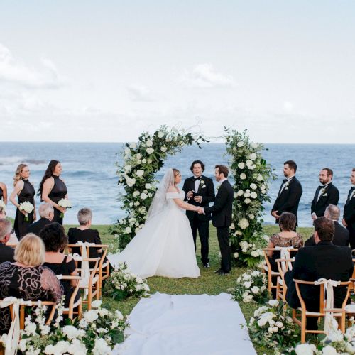 A wedding ceremony by the ocean, with a bride, groom, officiant, bridesmaids, and groomsmen, surrounded by flowers and guests seated in rows.