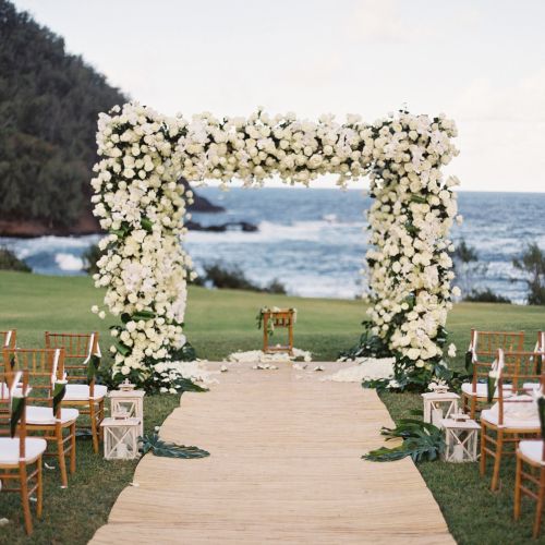 An outdoor wedding setup by the sea, with an aisle leading to a floral arch, surrounded by chairs, and a scenic ocean backdrop.