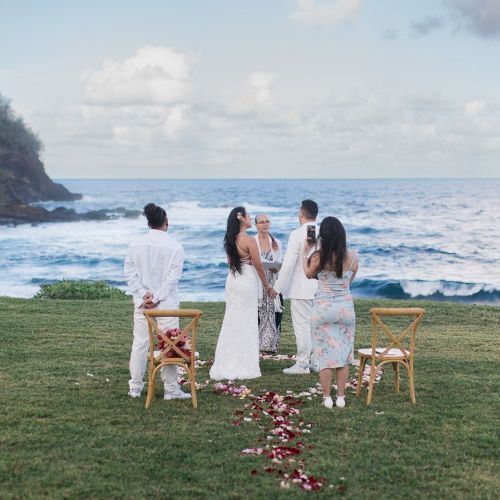 A small wedding ceremony is taking place near a coastline with waves in the background, featuring a few people standing and chairs with flower petals.