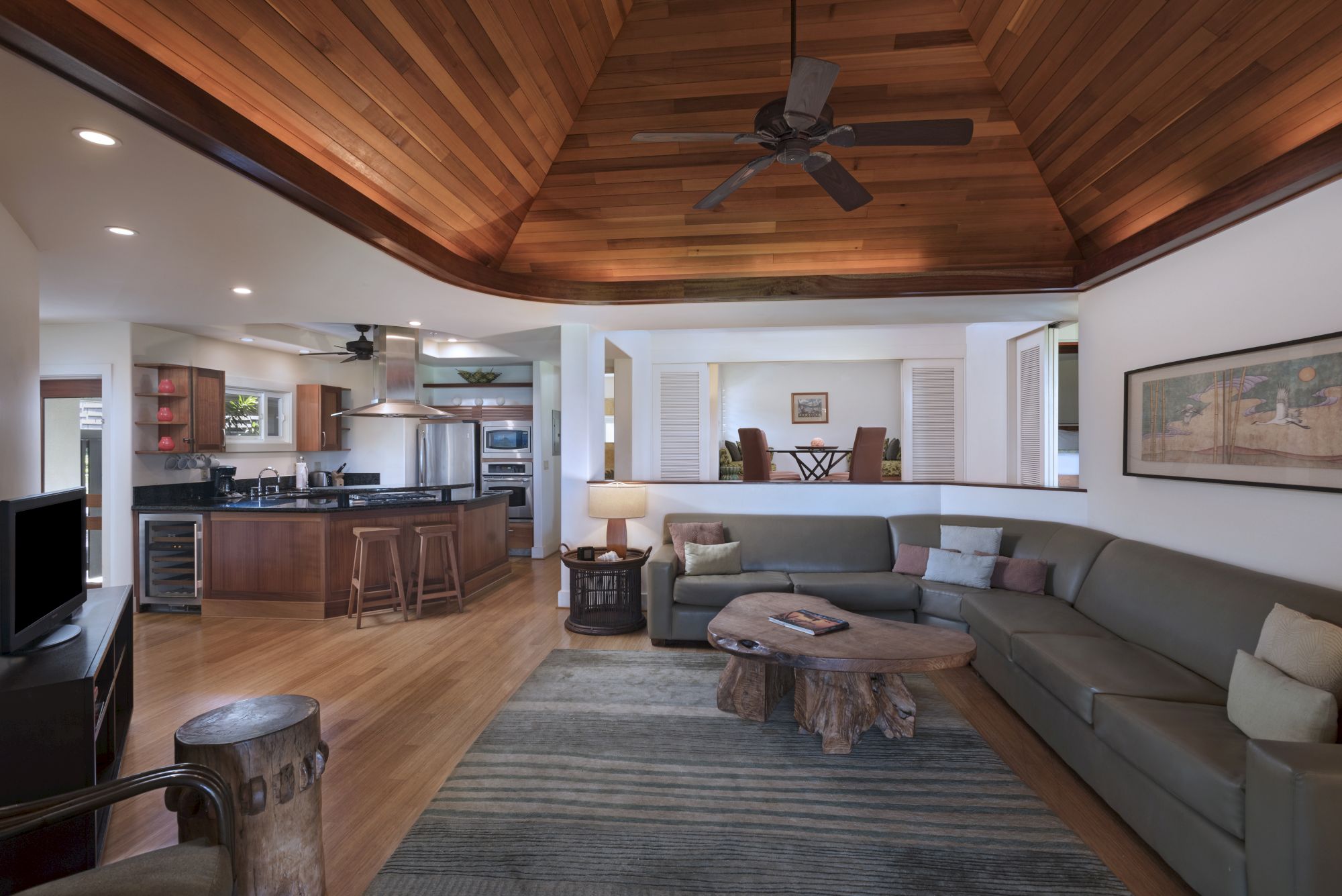 The image shows a stylish living room with a sectional sofa, rustic coffee table, ceiling fan, kitchen and dining area. The ceiling is wooden, adding warmth.