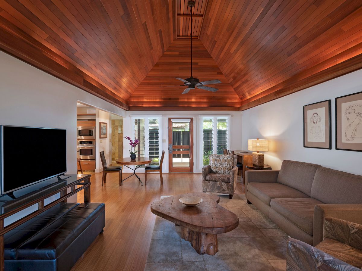 A cozy living room with a wooden ceiling, modern furniture, a TV, and artwork. It has a natural wood coffee table and warm lighting.