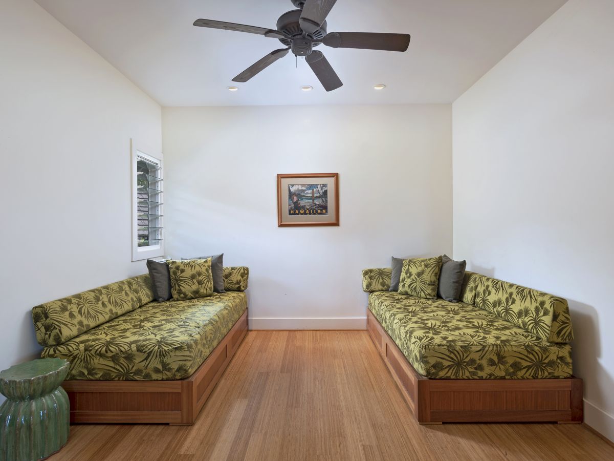 The image shows a room with two green-patterned daybeds, a ceiling fan, a framed picture on the wall, and wooden floors.