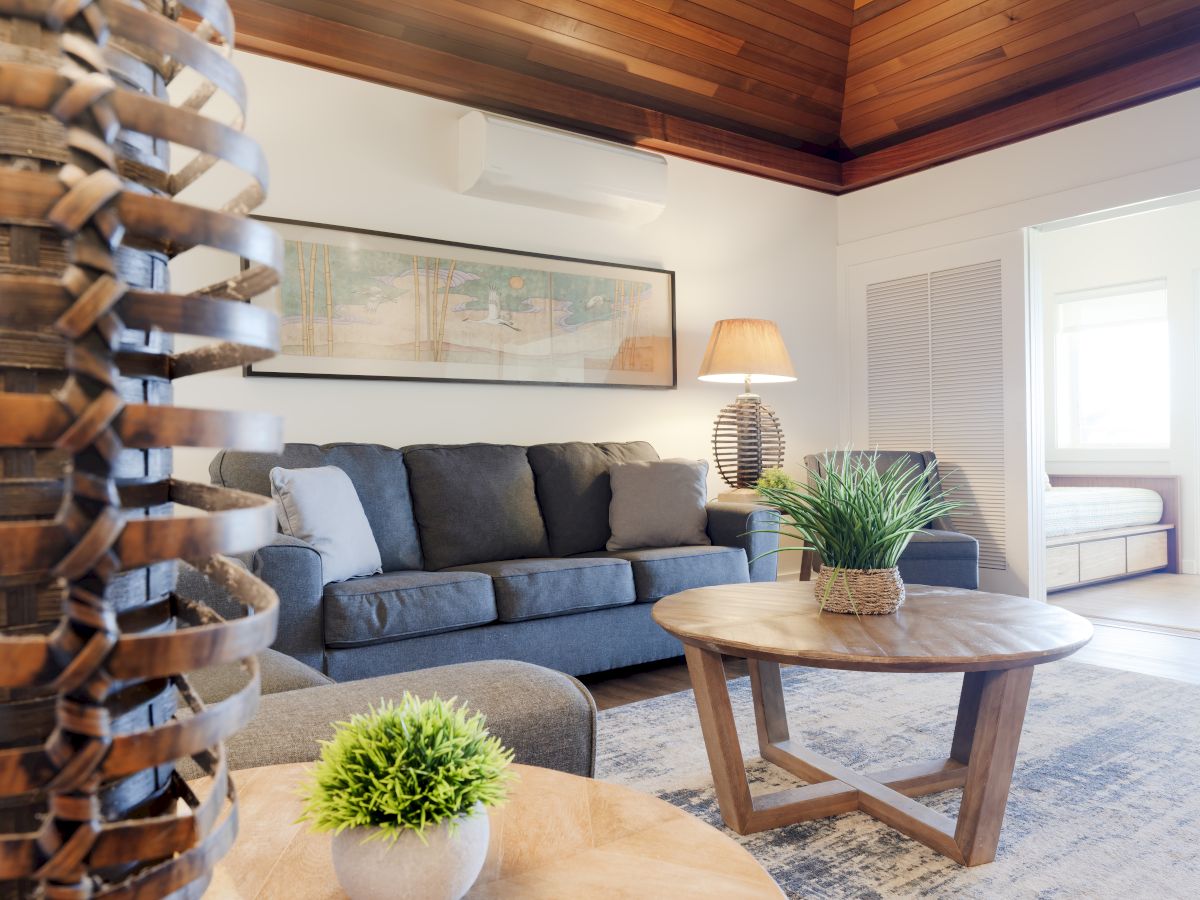 A cozy living room with a gray sofa, wooden coffee table, plants, and warm lighting, featuring a high wooden ceiling and wall artwork.