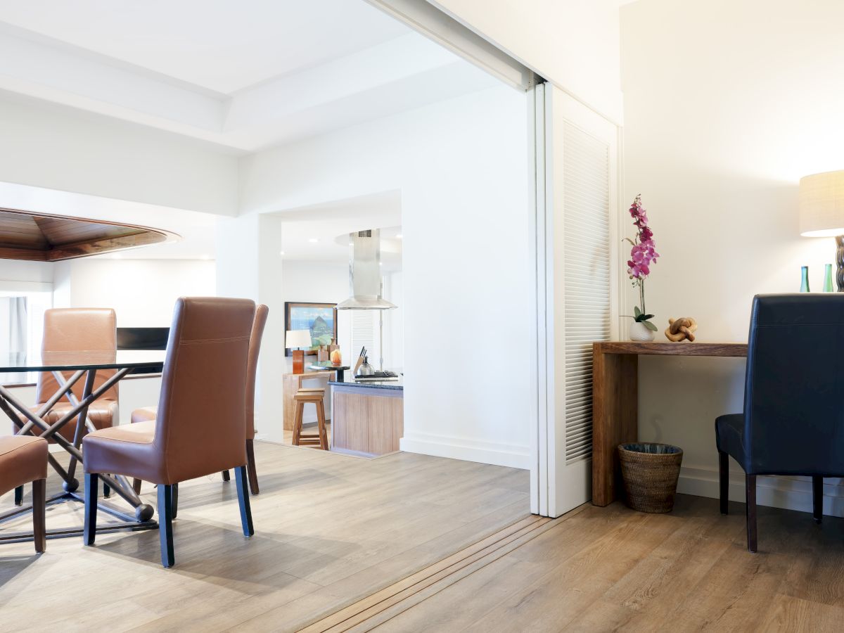 The image shows a modern, open dining and workspace area with brown chairs, wooden floors, and a small desk with a lamp and plant.