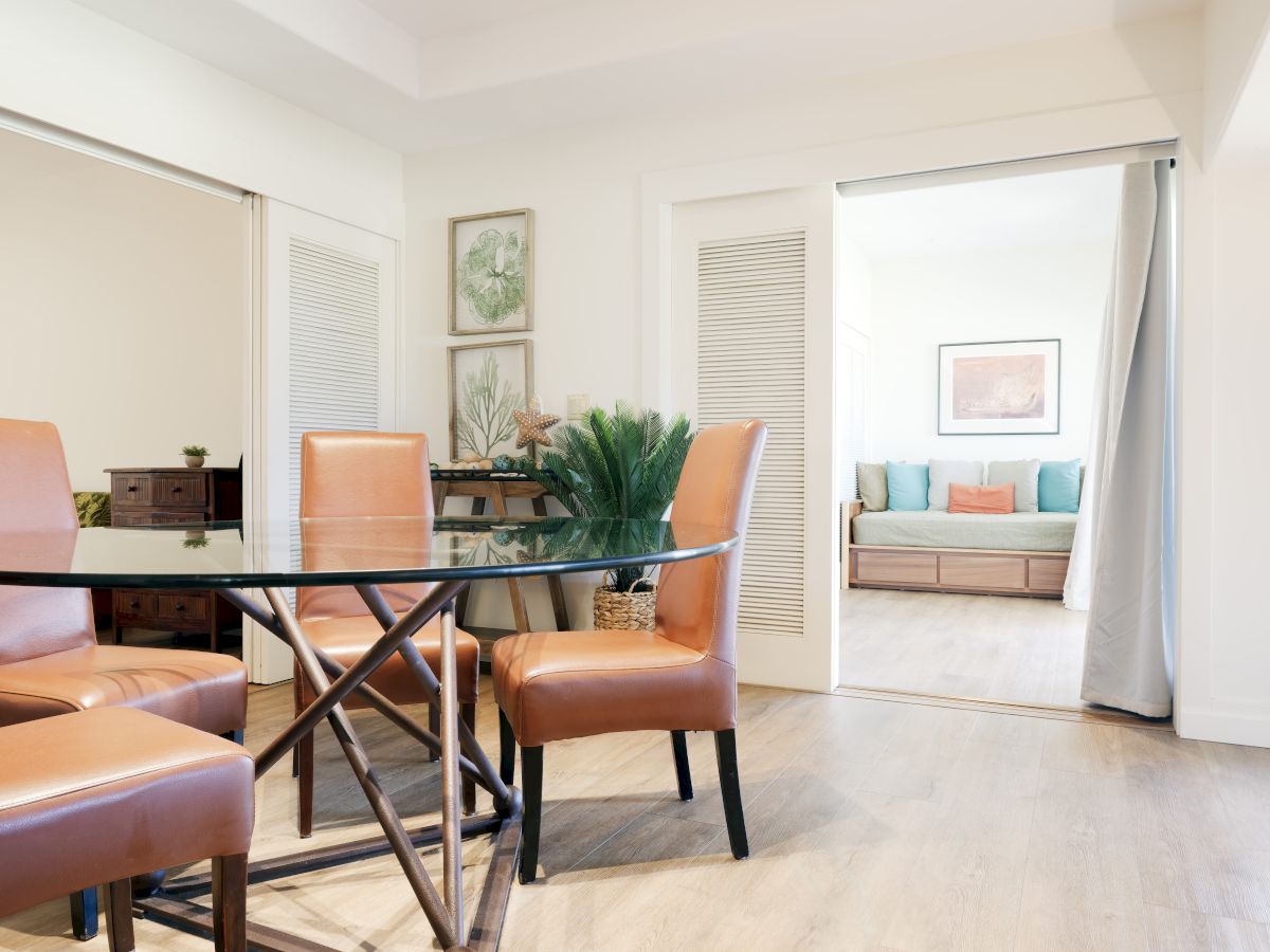 A dining area with tan chairs and a glass table, adjacent to a living space with a daybed and colorful cushions. Light and airy decor.