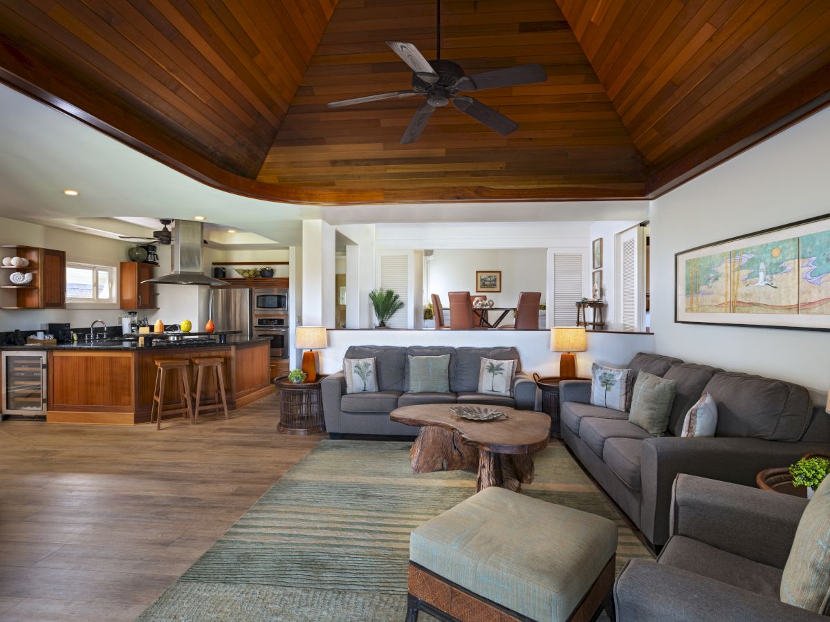 A spacious living room with a grey sectional sofa, wooden ceiling, open kitchen, artwork, and modern decor.