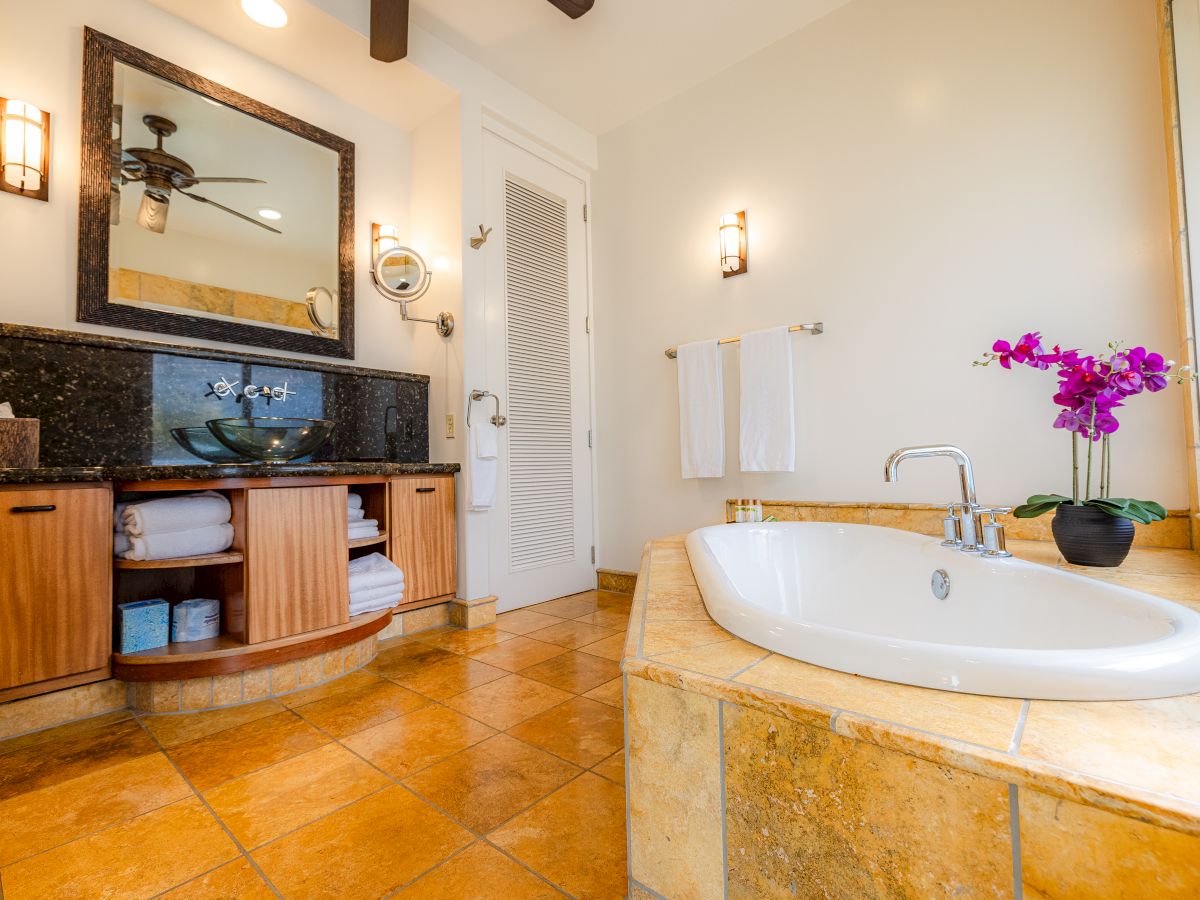 A bathroom with a corner tub, a wooden vanity, and a mirror. It features tiled flooring, white towels, and a small purple orchid on the tub ledge.