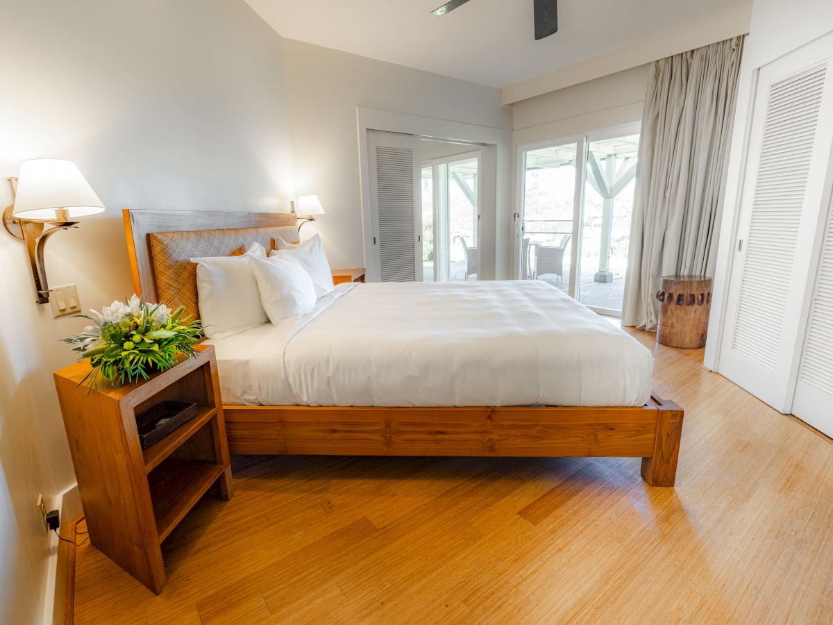 A cozy bedroom with a wooden bed, white linens, nightstand with flowers, and large windows with curtains, allowing natural light.