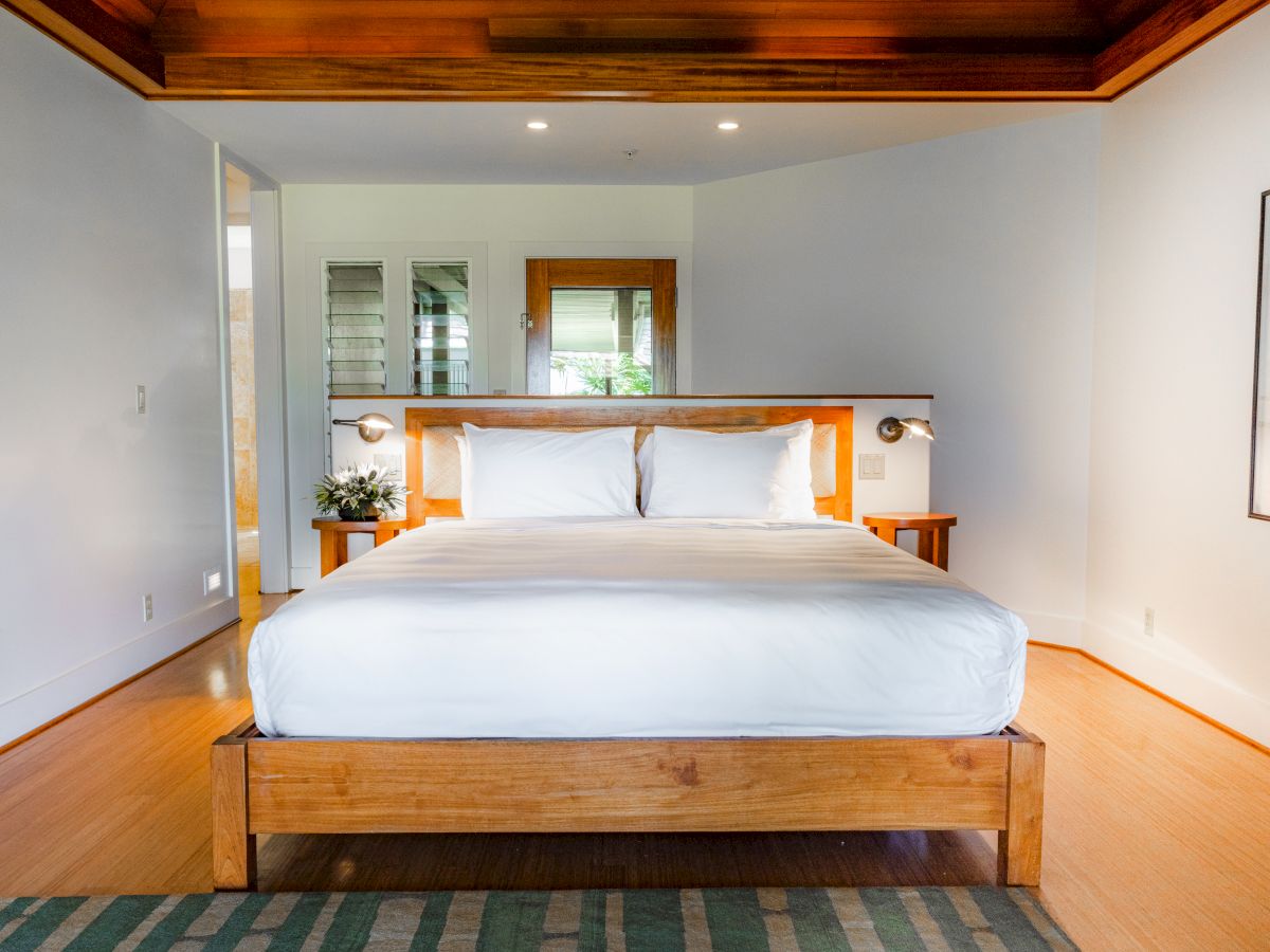 The image shows a modern bedroom with a wooden bed, white bedding, a rug, wall art, and a wooden ceiling feature, creating a cozy ambiance.