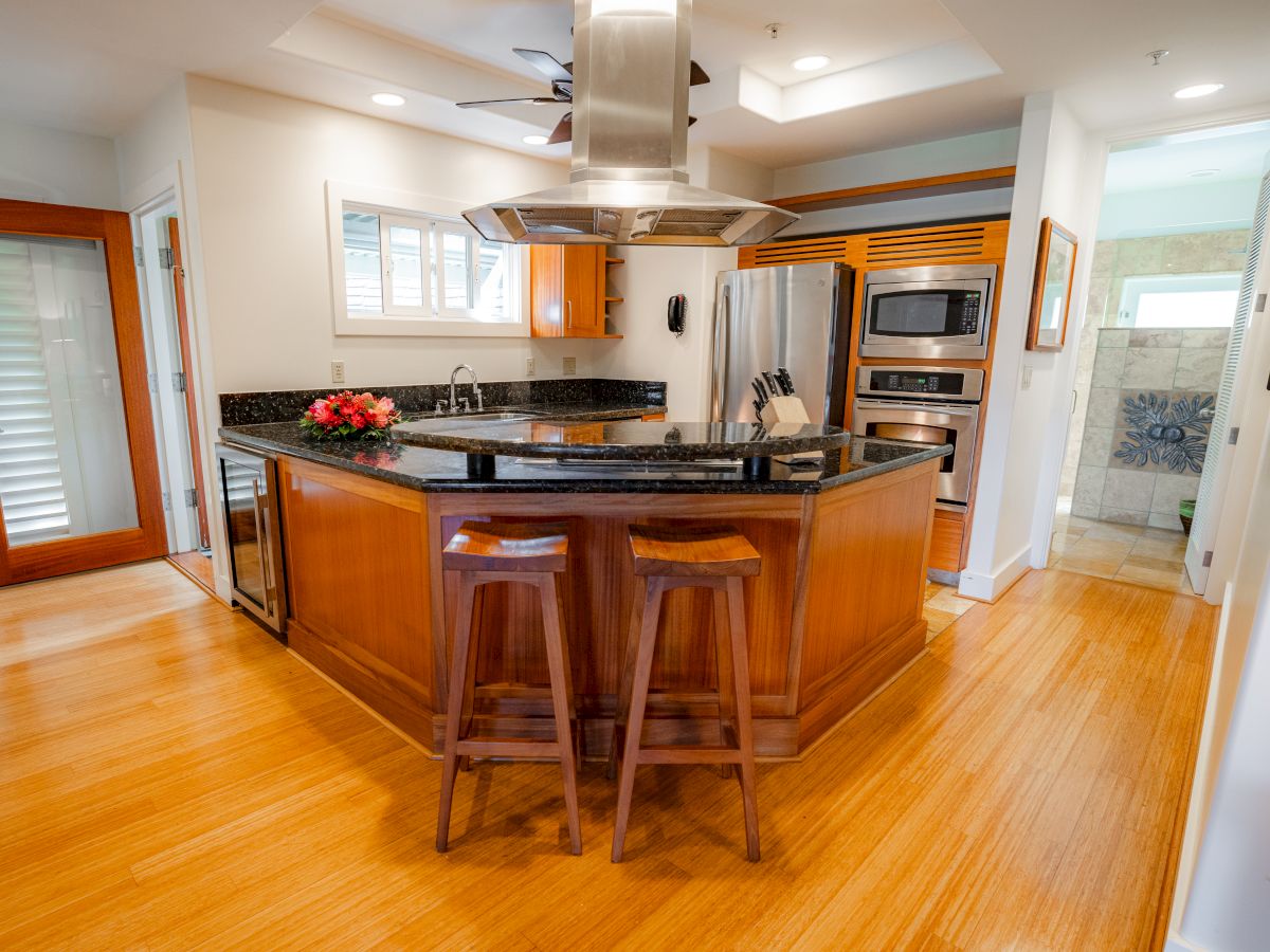 A modern kitchen with a wooden island, stools, stainless steel appliances, and a tiled floor. There are doors and windows providing light.