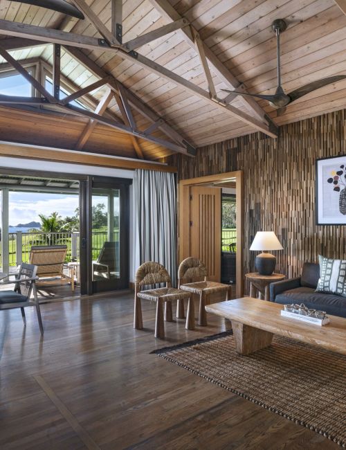 A cozy living room has wooden interiors, a seating area with a couch and tables, and large windows offering a scenic view of nature in the distance.