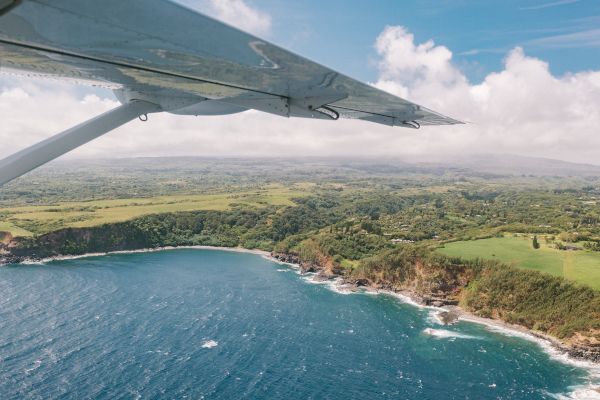 Aerial view from a plane wing over a coastline with blue ocean waves, green landscape, and a partly cloudy sky in the background.