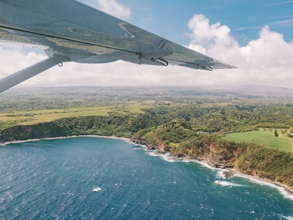 Aerial view from a plane wing over a coastline with blue ocean waves, green landscape, and a partly cloudy sky in the background.