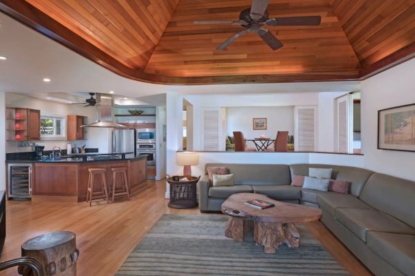 Spacious living area with a modern kitchen, sectional sofa, wooden ceiling, and a rustic coffee table. Open dining in the background.