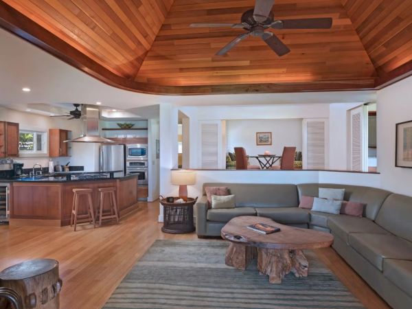Spacious living area with a modern kitchen, sectional sofa, wooden ceiling, and a rustic coffee table. Open dining in the background.
