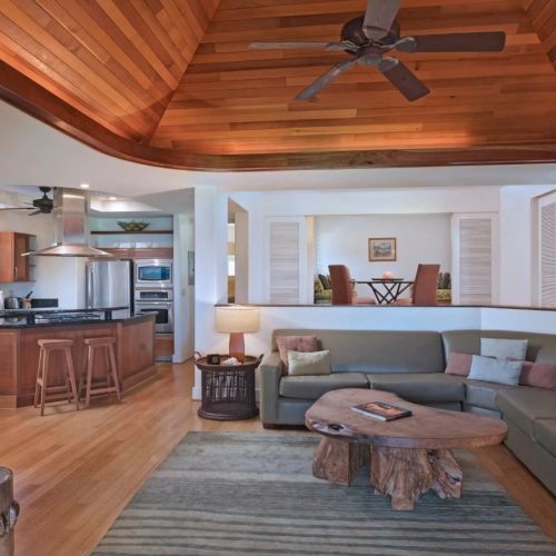 A spacious living area with a sectional sofa, rustic coffee table, open kitchen, wooden ceiling, and ceiling fan for a cozy ambiance.