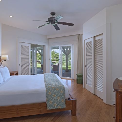 A cozy bedroom with a double bed, ceiling fan, sliding doors, and wooden furniture. Curtains and a view of a garden are visible.