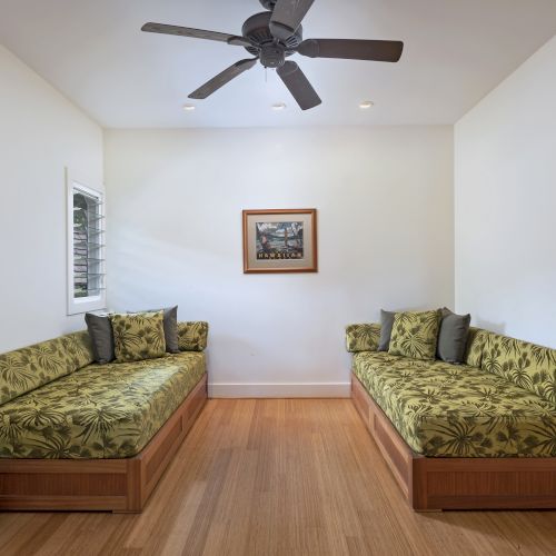 A room with two green floral sofas, a ceiling fan, a framed picture on the wall, and two stacked round tables on a wooden floor.