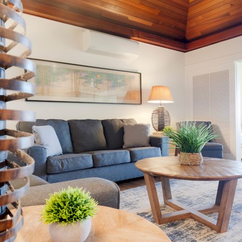 A cozy living room with a gray sofa, wooden coffee table, plants, and stylish decor. Warm lighting and wooden ceiling add a cozy touch.